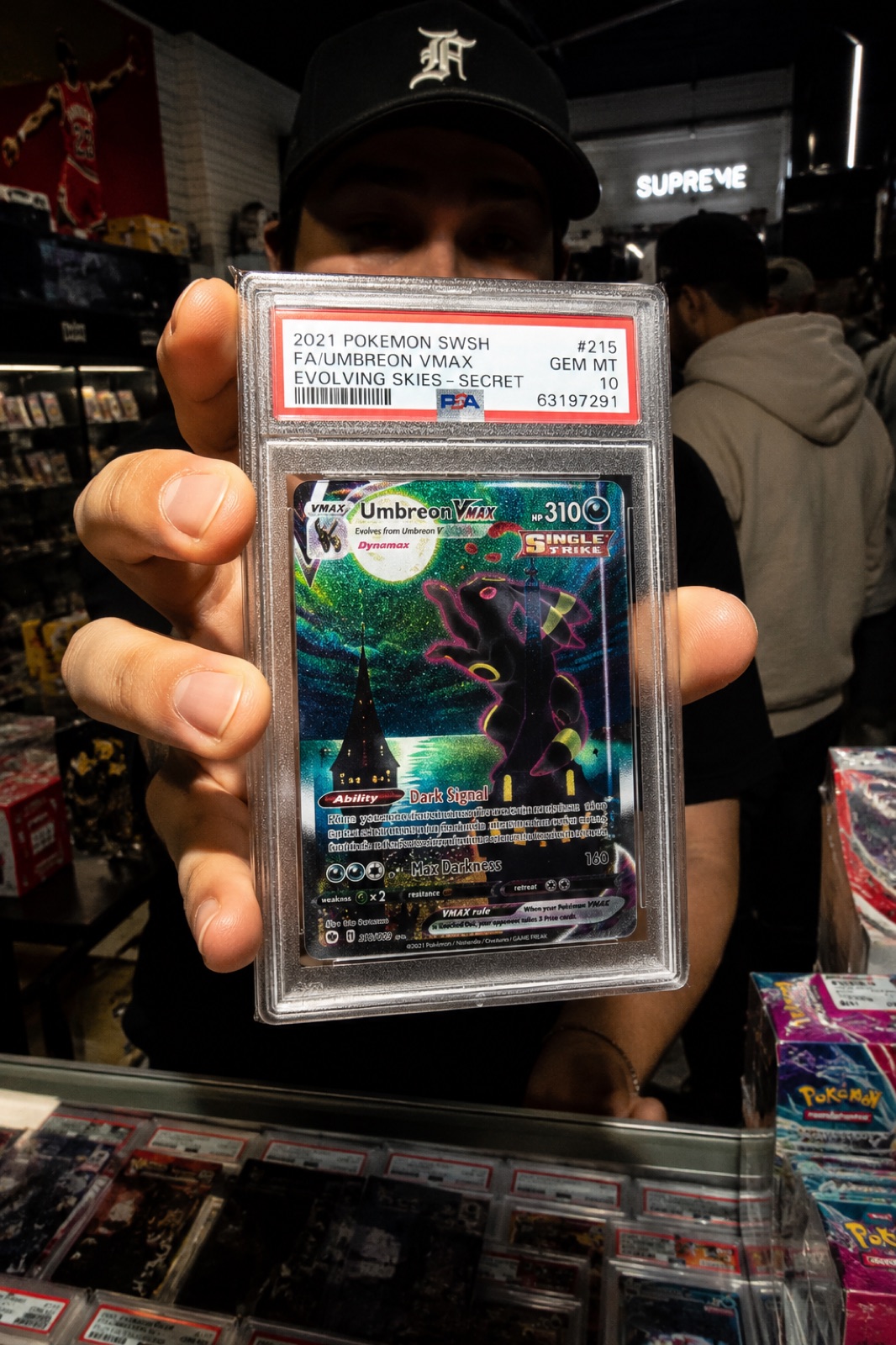 A collector at the Card Kennel counter holding a graded slab up to the camera, flash-lit